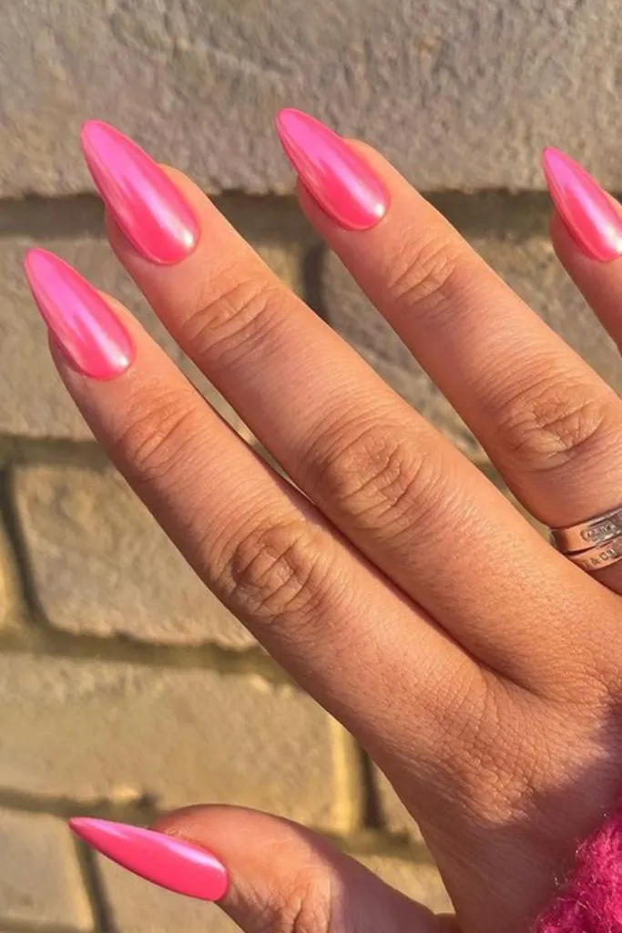 A hand with long, almond-shaped nails in a vibrant hot pink chrome finish, reflecting light.